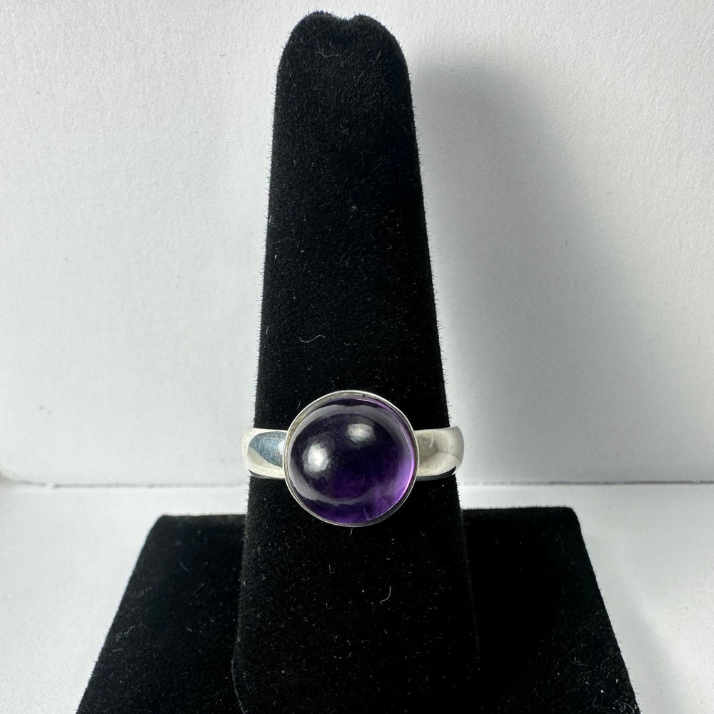 Silver ring with a round, translucent deep purple amethyst cabochon set in a smooth bezel, displayed on a black velvet ring holder.