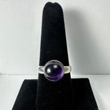 Silver ring with a round, translucent deep purple amethyst cabochon set in a smooth bezel, displayed on a black velvet ring holder.