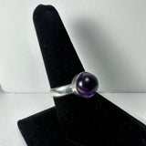 Silver ring with a polished round amethyst stone mounted on a smooth band, displayed on a black velvet ring holder against a white background.