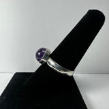 Silver ring with a polished round purple amethyst cabochon set in a bezel, displayed on a black velvet cone stand against a white background.