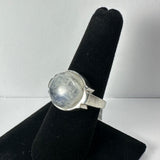 Silver ring with a large, round, translucent moonstone set in a simple bezel, displayed on a black velvet finger stand against a white background.