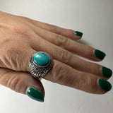 Hand with dark green nail polish wearing a size 7 ring featuring a smooth oval turquoise stone set in an intricately patterned silver band.