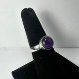 Silver ring with a smooth, rounded amethyst stone set in a bezel setting, displayed on a black velvet ring holder against a white background.