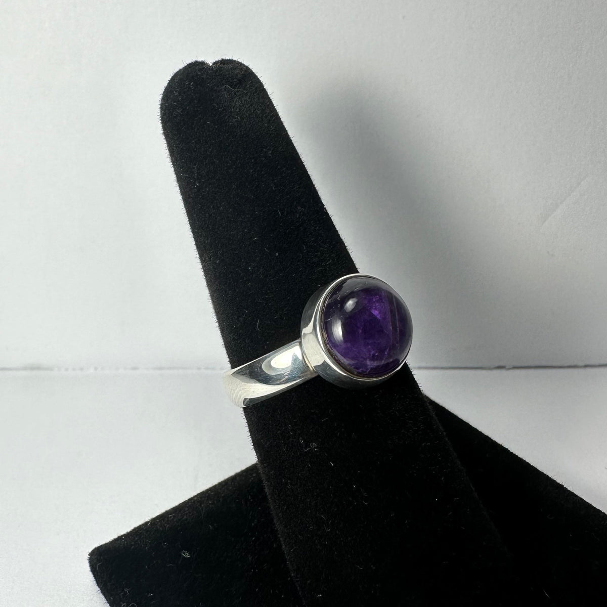 Silver ring with a smooth, rounded amethyst stone set in a bezel setting, displayed on a black velvet ring holder against a white background.