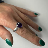 Hand with green nail polish wearing a silver ring featuring a round, polished purple amethyst stone on the middle finger, shown against a white background.