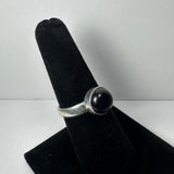 Silver ring with a round, polished dark purple amethyst stone set in a bezel on a black velvet ring holder against a plain white background.