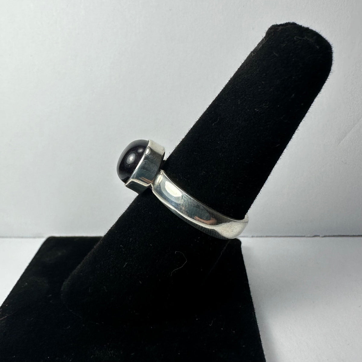Silver ring with a round dark cabochon gemstone mounted on top, displayed on a black velvet ring holder against a white background.