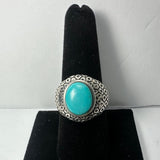 Silver ring with an oval turquoise stone set in the center, featuring a patterned band, displayed on a black velvet ring holder against a white background.