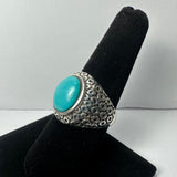Silver ring with a hexagonal-patterned band featuring a smooth, oval turquoise stone set in the center, displayed on a black velvet ring holder.