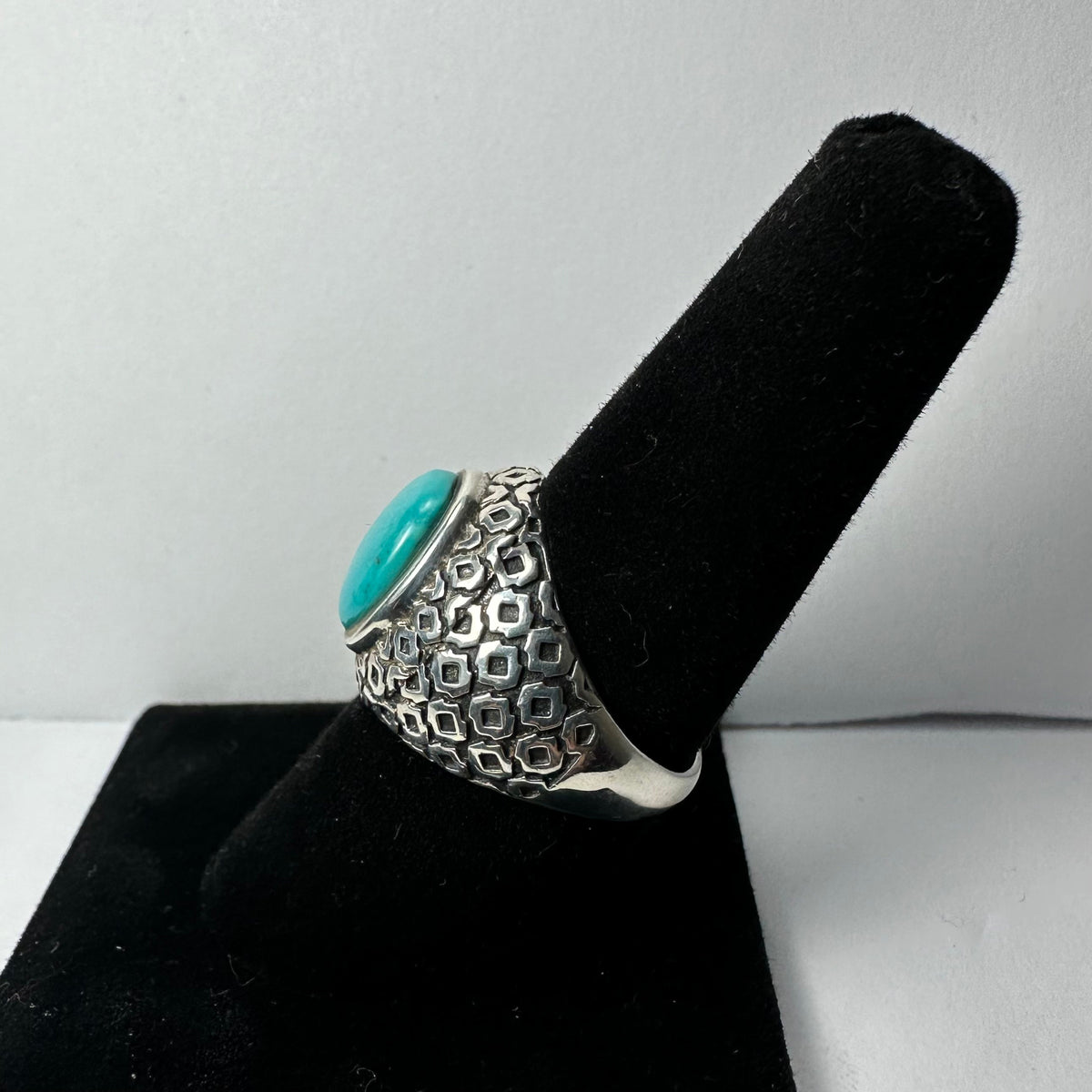 Silver ring with a round turquoise stone centered on a patterned band, displayed on a black ring holder against a light background.