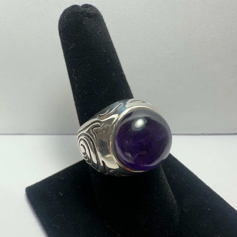 Silver ring with carved swirling patterns and a large polished deep purple amethyst cabochon, displayed on a black velvet finger stand against a plain white background.