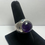 Silver ring with carved swirling patterns and a large polished deep purple amethyst cabochon, displayed on a black velvet finger stand against a plain white background.