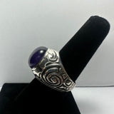 Silver ring with intricate swirling patterns and a polished oval amethyst stone set in the center, displayed on a black velvet ring holder.