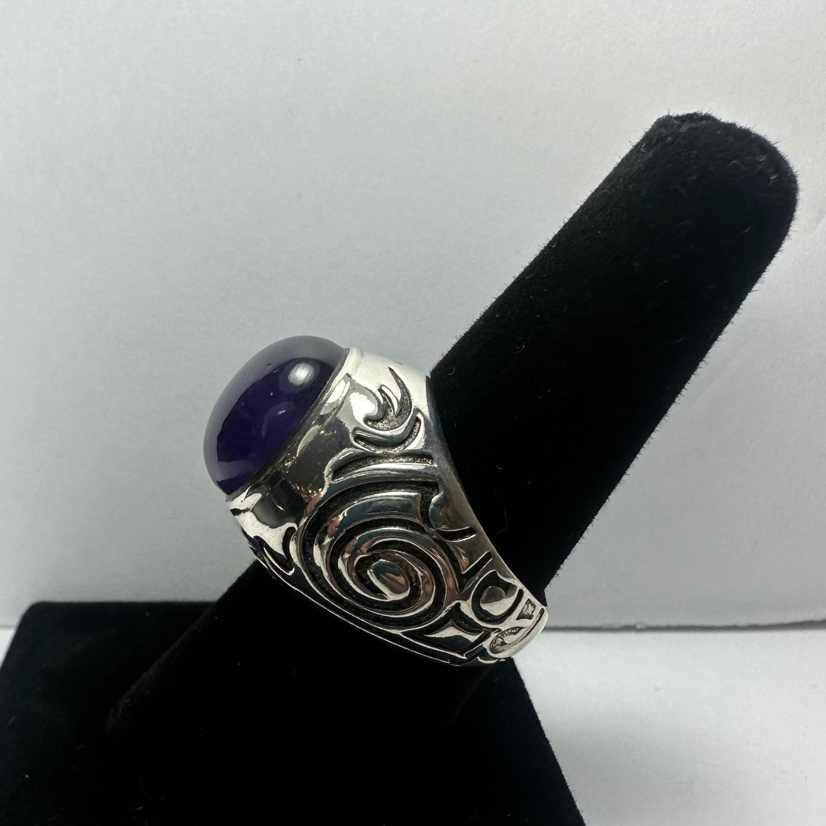 Silver ring with intricate swirling patterns and a polished oval amethyst stone set in the center, displayed on a black velvet ring holder.