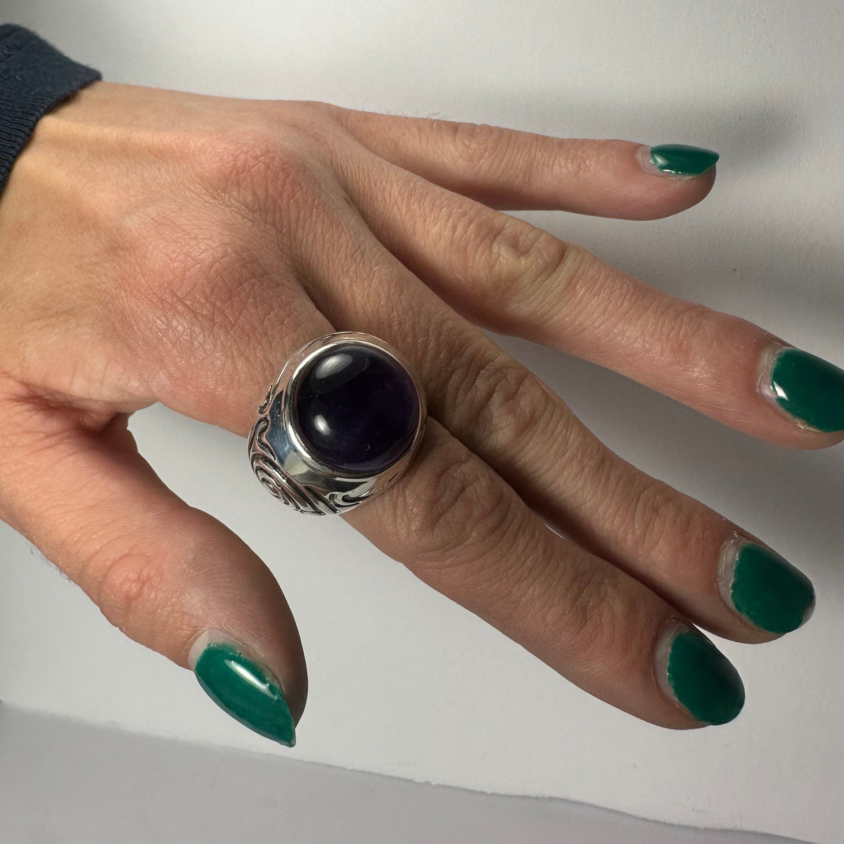 Hand with green nail polish wearing a silver ring featuring a large, polished, dark purple amethyst stone and engraved leaf patterns on the band.