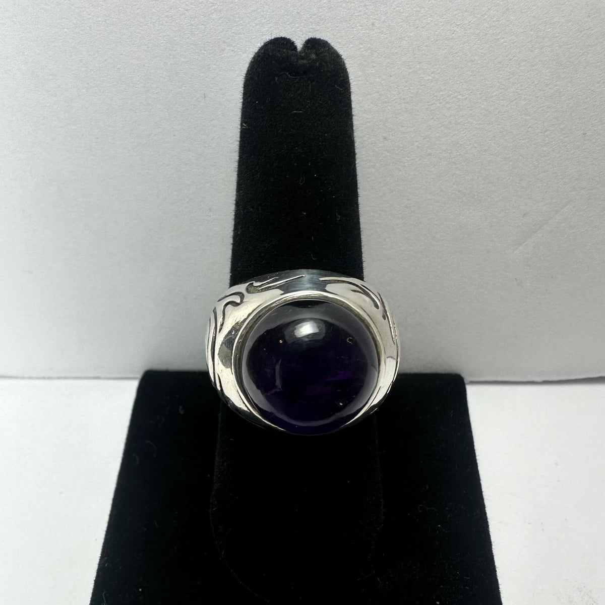 Silver ring with a large round deep purple amethyst stone set in a smooth bezel, displayed upright on a black velvet ring stand.