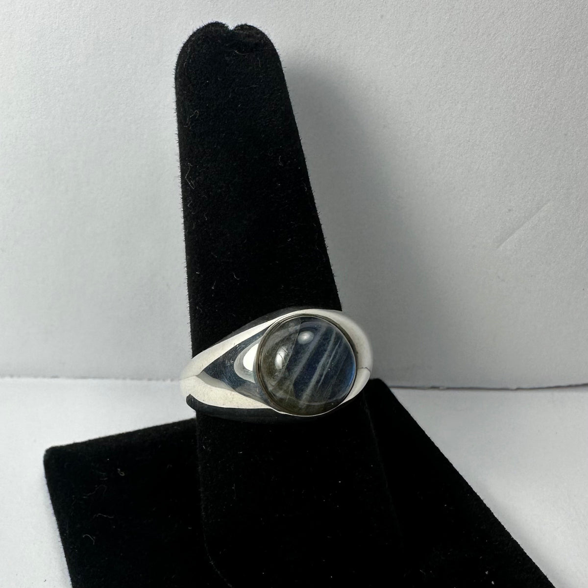 Silver ring with a polished round labradorite stone featuring blue and gray hues, displayed on a black velvet finger stand against a plain gray background.