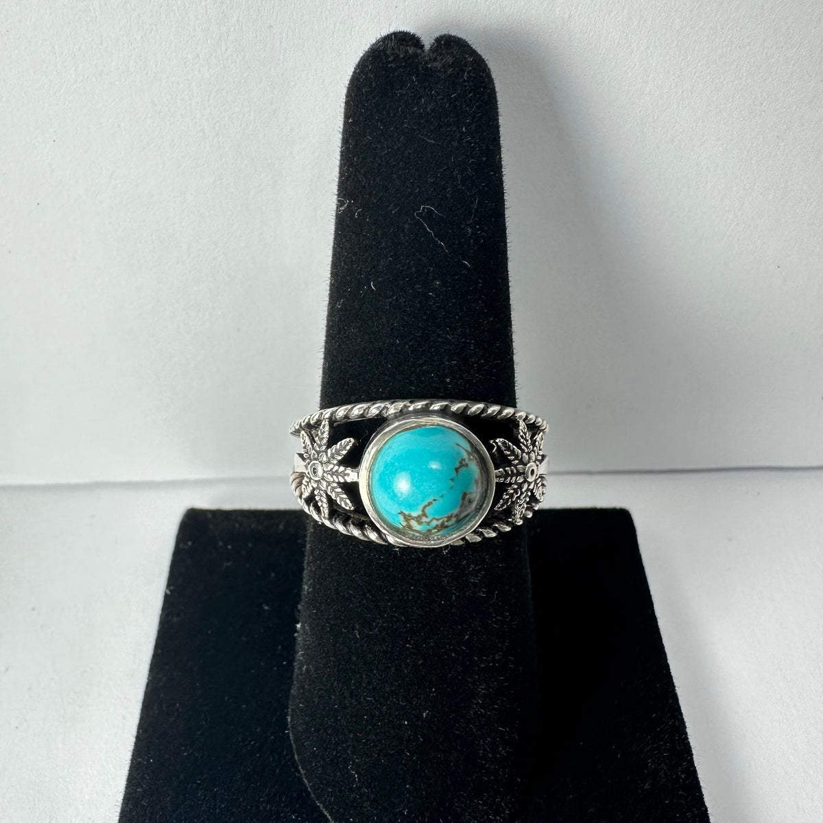 Silver ring with a round turquoise stone center and floral accents on a black velvet ring display stand.
