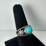 Silver ring featuring a round turquoise stone with black matrix set beside a detailed silver leaf motif on a black velvet ring stand.
