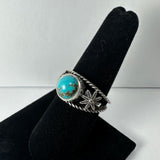 Silver ring with round turquoise stone center and adjacent decorative star-shaped design on textured black display stand