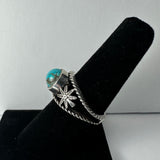 Silver ring featuring a round turquoise stone and a detailed silver starburst design on a twisted band, displayed on a black velvet ring holder.