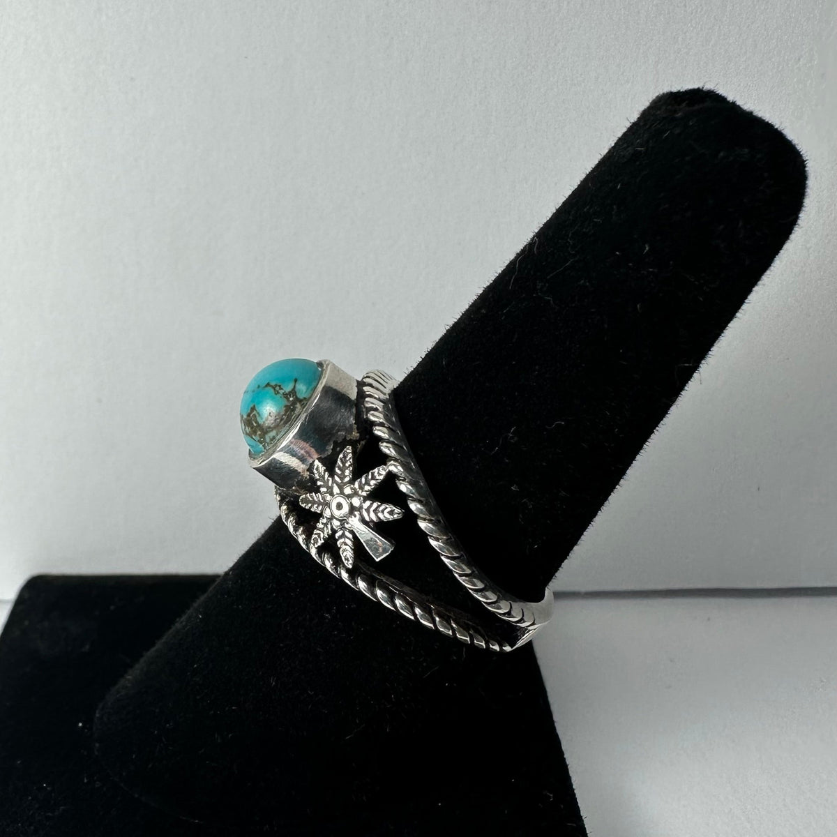 Silver ring featuring a round turquoise stone and a detailed silver starburst design on a twisted band, displayed on a black velvet ring holder.