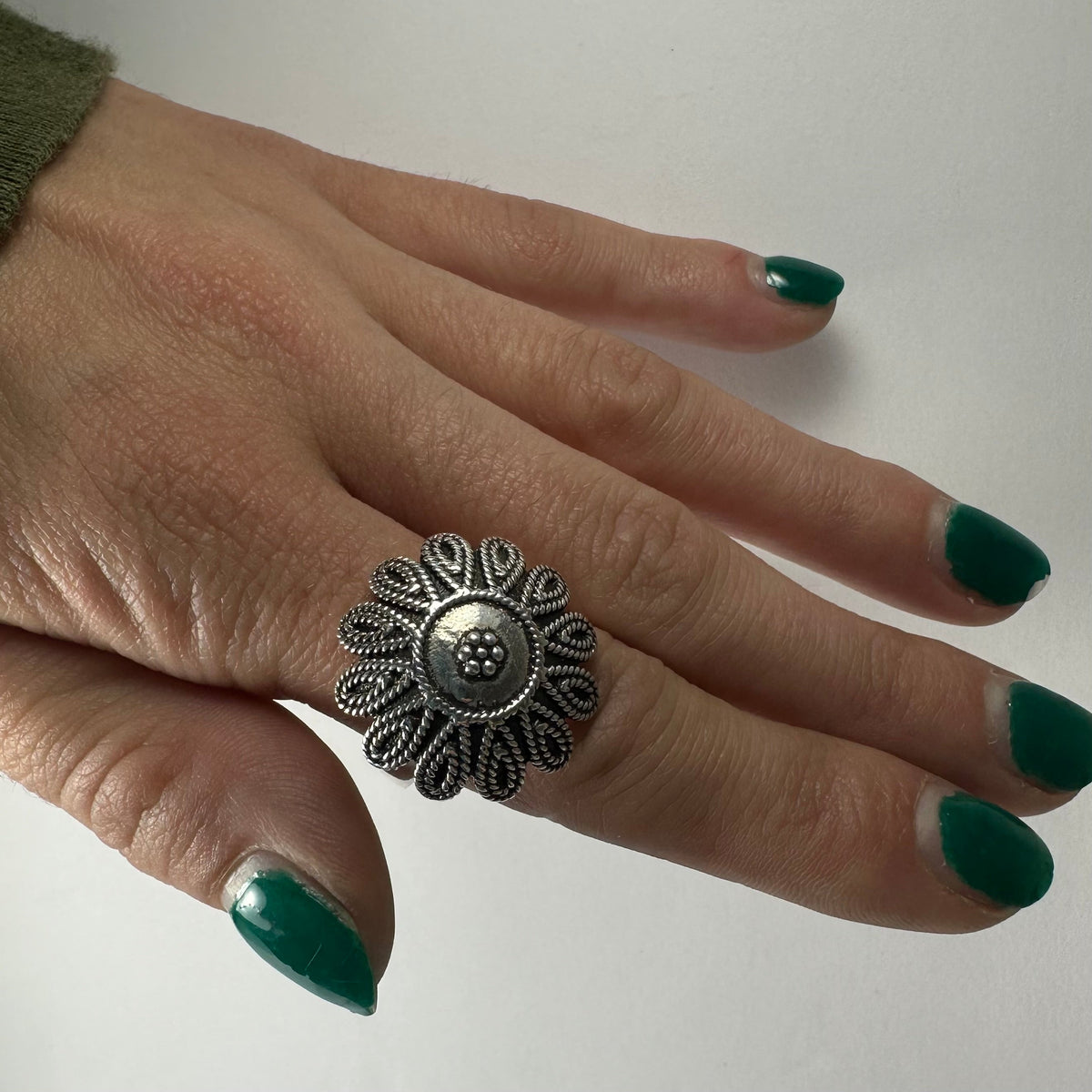Silver ring shaped like a detailed flower with textured petals, worn on a finger with dark green nail polish against a plain background.