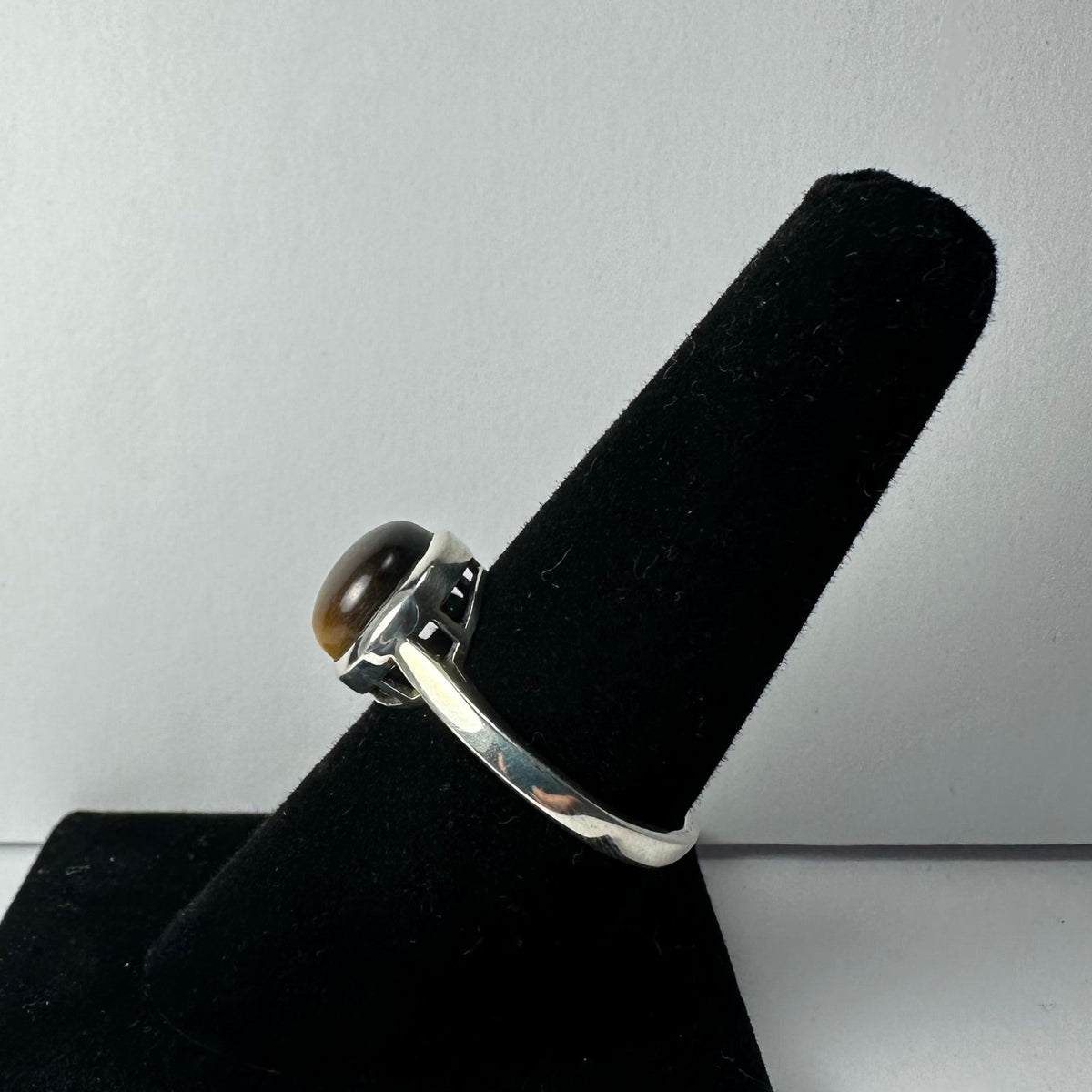 Silver ring with a round, polished brown tiger's eye stone set in a bezel, shown angled on a black velvet ring holder against a gray background.