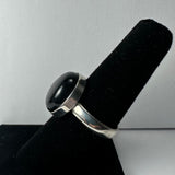 Silver band ring featuring a polished oval black onyx stone, displayed on a black velvet ring stand against a light gray background.