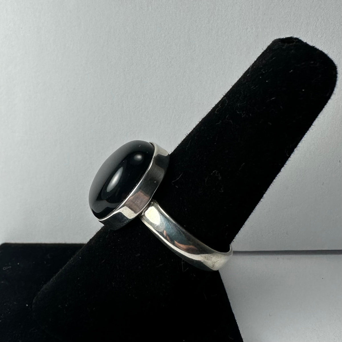Silver band ring featuring a polished oval black onyx stone, displayed on a black velvet ring stand against a light gray background.