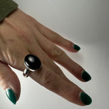 Hand with green polished nails wearing a silver ring featuring a smooth, oval black onyx stone on the middle finger against a plain light background.
