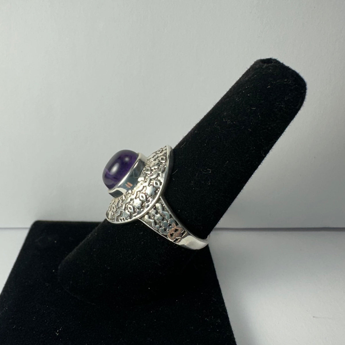 Silver ring with a round cabochon amethyst stone set in a wide hammered and engraved band, displayed on a black velvet ring holder against a plain background.