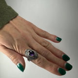 Hand with dark green nail polish wearing a silver ring featuring a smooth, oval-shaped purple amethyst stone, shown against a plain light background.