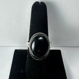Silver ring featuring a large polished oval black onyx stone set on a black velvet display stand against a plain white background.