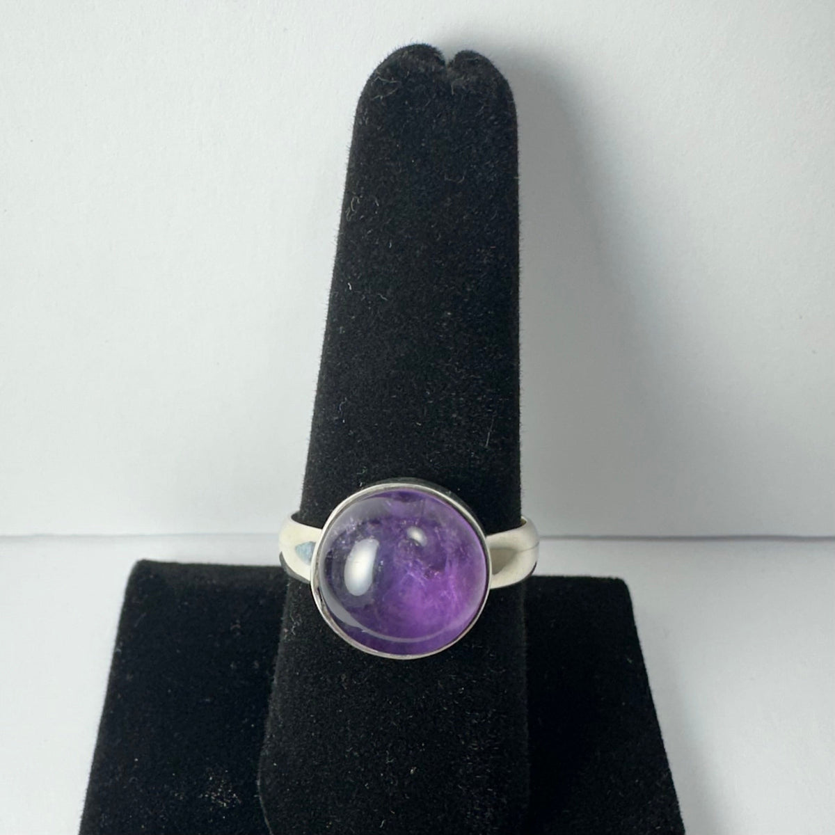 Silver ring with a smooth round amethyst stone set in a simple bezel, displayed upright on a black velvet ring holder against a white background.