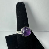 Silver ring with a polished round amethyst stone set in a simple bezel, displayed upright on a black velvet ring stand against a light gray background.