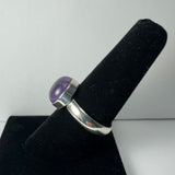 Silver ring with a rounded purple amethyst stone set in a bezel, displayed upright on a black velvet ring holder against a light background.