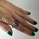 Hand with green nail polish wearing a silver ring featuring a polished round amethyst stone, shown against a plain white background.