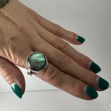 Hand with green painted nails wearing a silver ring featuring a large, round labradorite stone with blue-green iridescence.