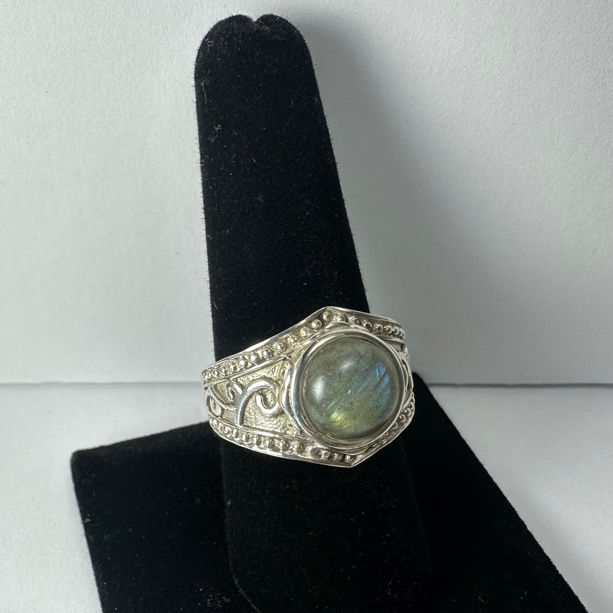 Silver ring with ornate filigree details and a round, polished labradorite stone displaying blue and green flashes, shown on a black velvet stand.