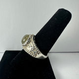 Silver ring with an oval labradorite stone featuring subtle green and blue hues and ornate scrollwork details on the band, displayed on a black velvet ring holder.