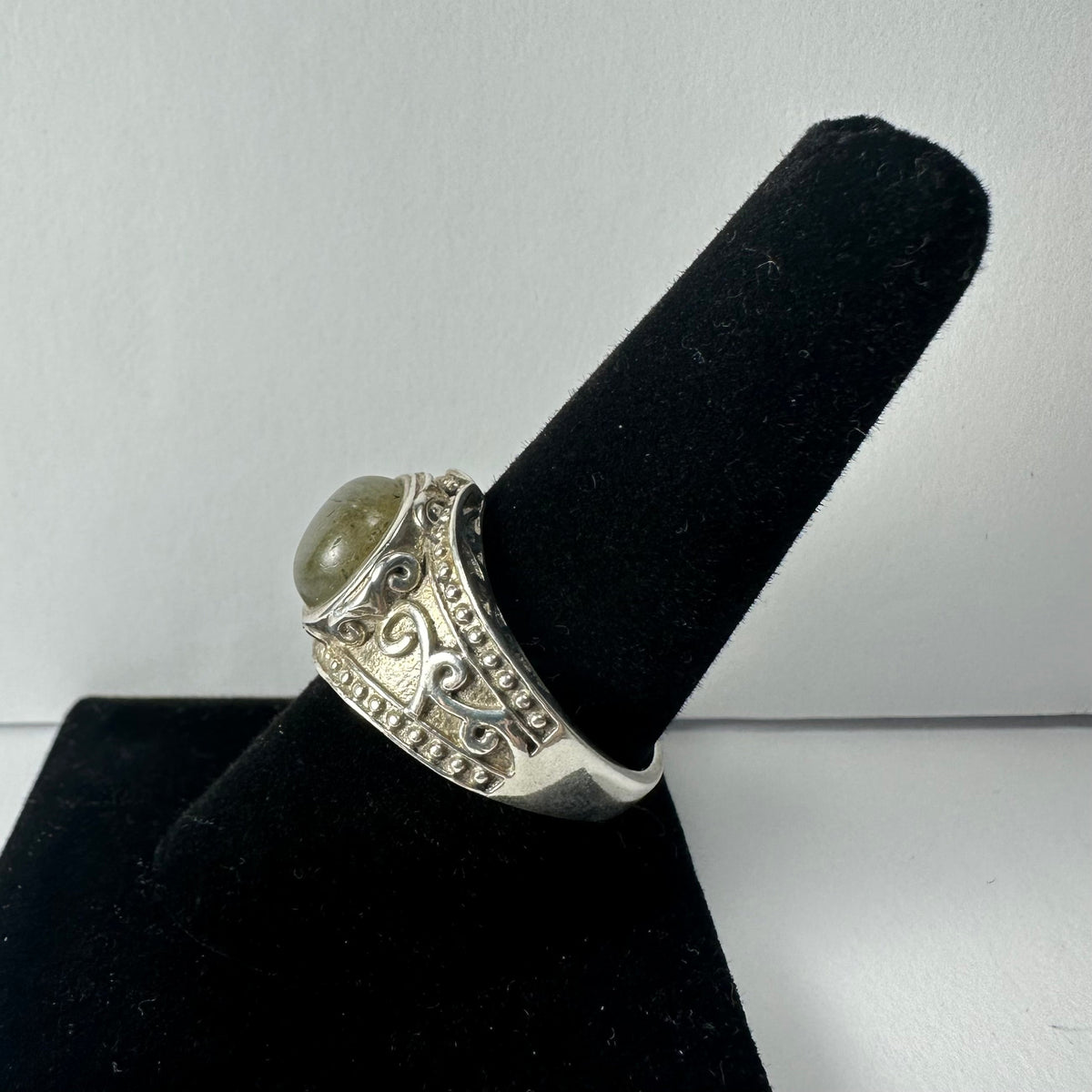 Silver ring with an oval labradorite stone featuring subtle green and blue hues and ornate scrollwork details on the band, displayed on a black velvet ring holder.