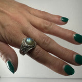 Hand with green nail polish wearing a silver ring featuring a round, iridescent labradorite stone and intricate engraved detailing on the band.