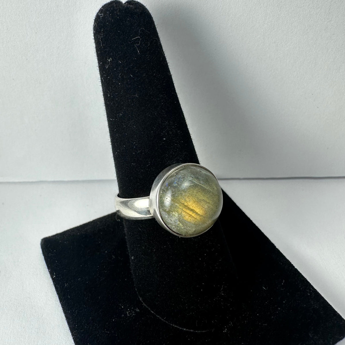 Silver ring with round labradorite gemstone displaying iridescent blue and yellow flashes, shown on black velvet ring stand against white background.