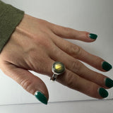 Hand with green nail polish wearing a silver ring featuring a round labradorite stone with yellow and blue iridescence, shown on index finger against a plain background.
