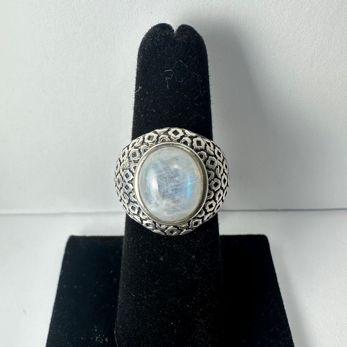Round moonstone cabochon set in an ornate silver band with floral and geometric patterns, displayed on a black velvet ring holder.