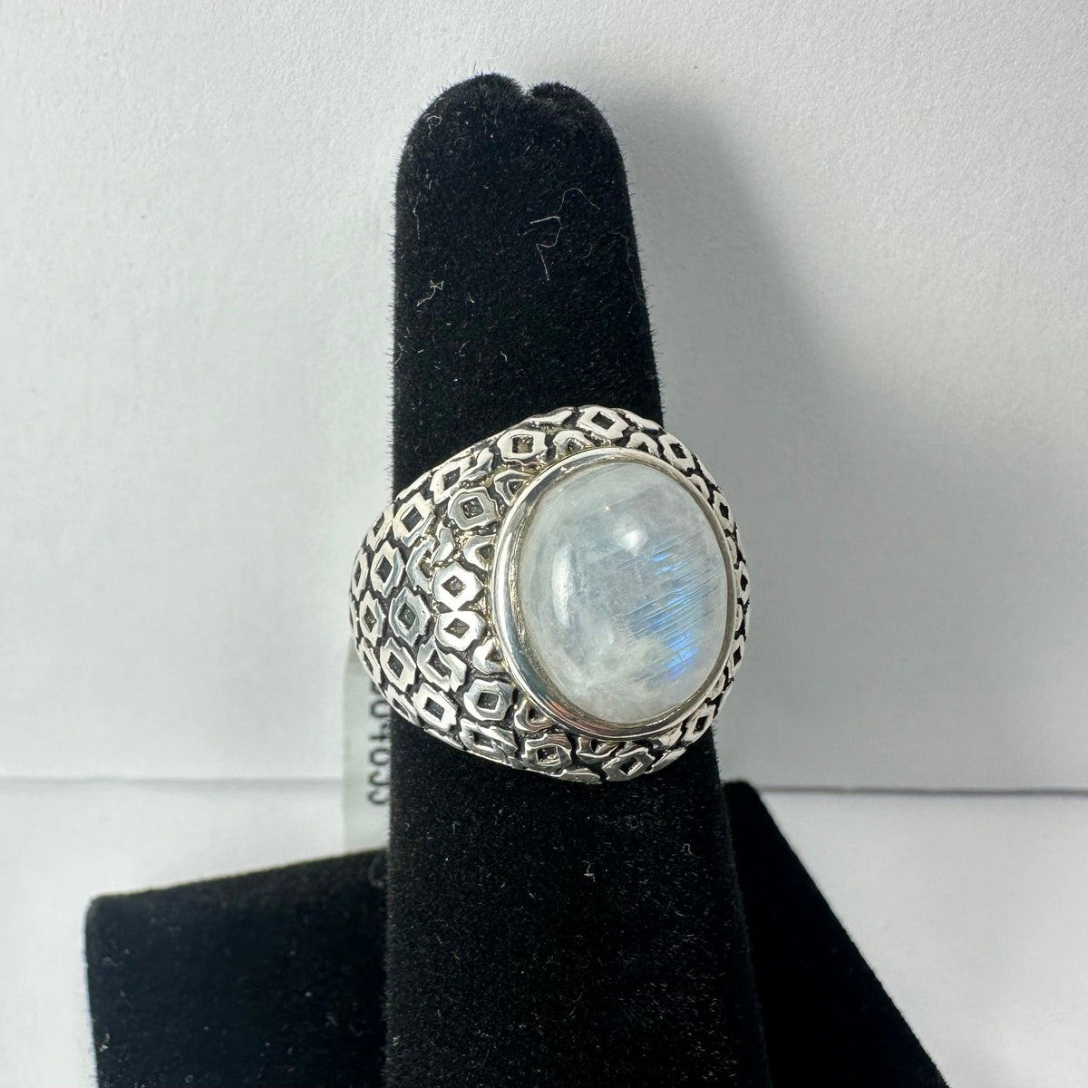 Silver ring with an oval moonstone center featuring a subtle iridescent glow and an intricate geometric pattern on the band, displayed on a black velvet ring holder.