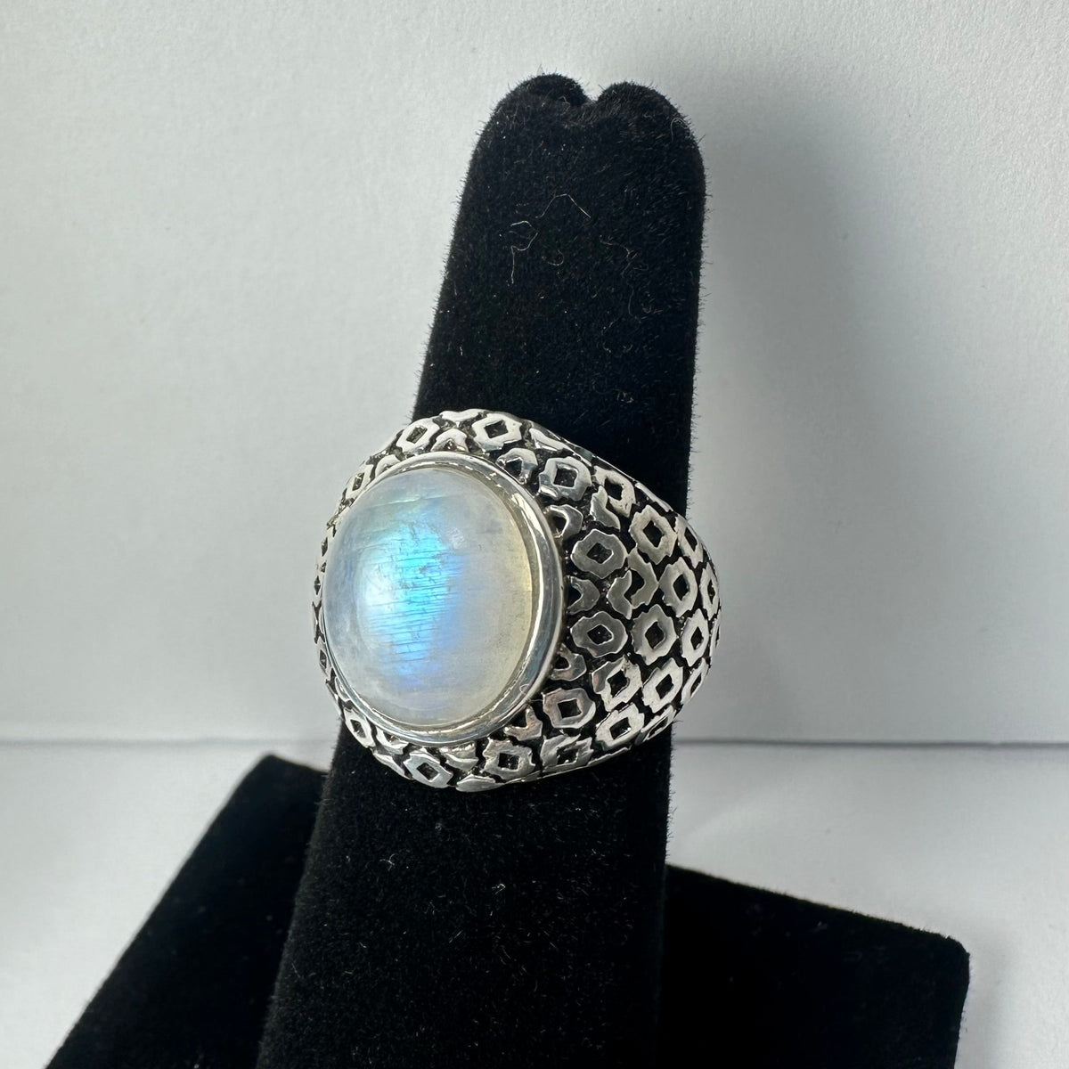 Silver ring with an oval moonstone featuring a blue sheen, set in an intricately patterned silver band, displayed on a black velvet stand.