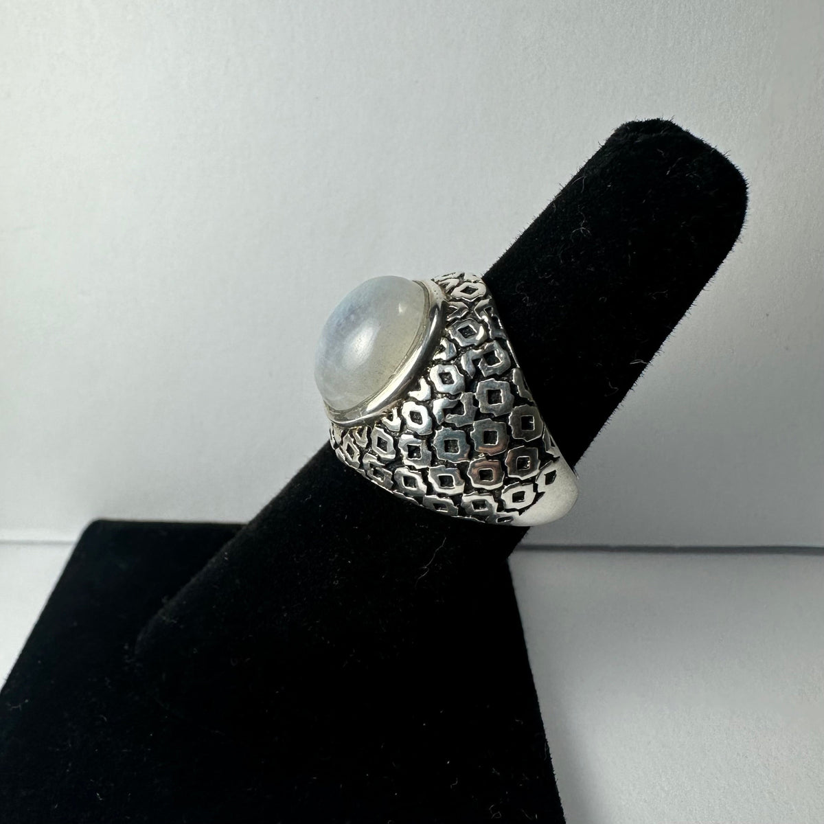 Silver ring with a raised moonstone cabochon center and an engraved geometric pattern on the wide band, displayed on a black ring holder.
