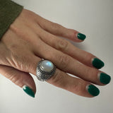 Hand with green nail polish wearing an ornate silver ring featuring a round, translucent white moonstone in the center, shown on a plain light background.